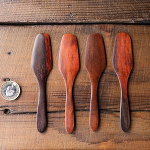 Può includere: Quattro spatole di legno con diversi motivi di venatura del legno. Le spatole sono disposte in fila su una superficie di legno.