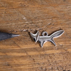 May include: A silver-toned fox-shaped charm featuring a smooth, reflective surface and a small circular loop at the top for attachment. The charm is positioned next to the tip of a grey pencil to provide a sense of scale for the small metal object.