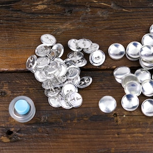 May include: A pile of silver metal snap buttons on a wooden surface. There is a blue plastic button press tool in the bottom left corner of the image.