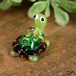 Könnte beinhalten: Eine kleine grüne Glas-Schildkrötenfigur mit braunen Schalen-Details. Die Schildkröte hat große schwarze Augen und einen grünen Kopf.