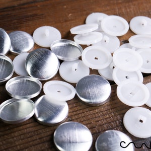 Puede incluir: Una colección de botones redondos plateados y respaldos de botones blancos dispersos sobre una superficie de madera. Los botones tienen un acabado de metal cepillado. Los respaldos blancos son de plástico.