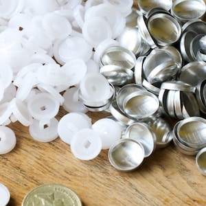 Puede incluir: Un montón de botones a presión de plástico blanco y un montón de botones a presión de metal plateado. Una moneda británica está en primer plano.