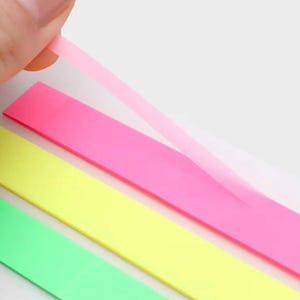May include: A set of three rectangular adhesive page markers in neon pink, neon yellow, and neon green. The top neon pink marker is partially peeled back, revealing a translucent, thin, and flexible plastic material used for indexing.
