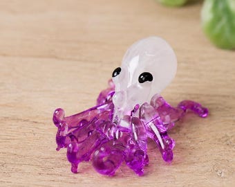 May include: A small glass figurine of an octopus with a translucent white head and deep purple tentacles. The octopus has two small black eyes and is sculpted with multiple curved arms extending outward from the base of its rounded head.