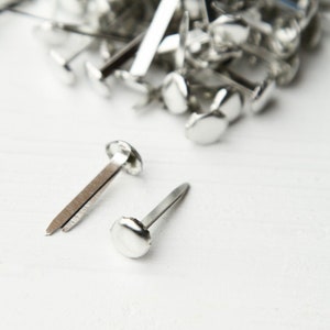 Op de afbeelding: Close-up van zilveren metalen bevestigingsmiddelen, ook wel paper fasteners of brads genoemd, verspreid op een wit oppervlak. Twee bevestigingsmiddelen zijn scherp, met hun ronde koppen en gespleten pinnen. De bevestigingsmiddelen worden gebruikt voor het vastzetten van papier of andere materialen.