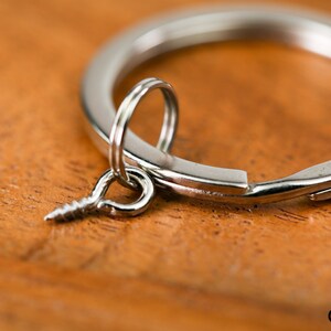 May include: A silver key ring with a screw-shaped charm attached to it. The key ring is lying on a wooden surface.