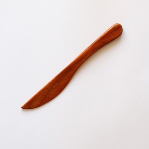 May include: A handcrafted wooden butter knife with a curved blade and rounded handle. The knife is made of reddish-brown wood and is set against a white background. This kitchen utensil is ideal for spreading butter or soft cheeses.