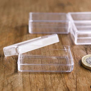 May include: Clear, rectangular plastic boxes with hinged lids, ideal for small items. One box is open, lid ajar. A British one-pound coin is next to the boxes. The boxes are on a wooden surface.