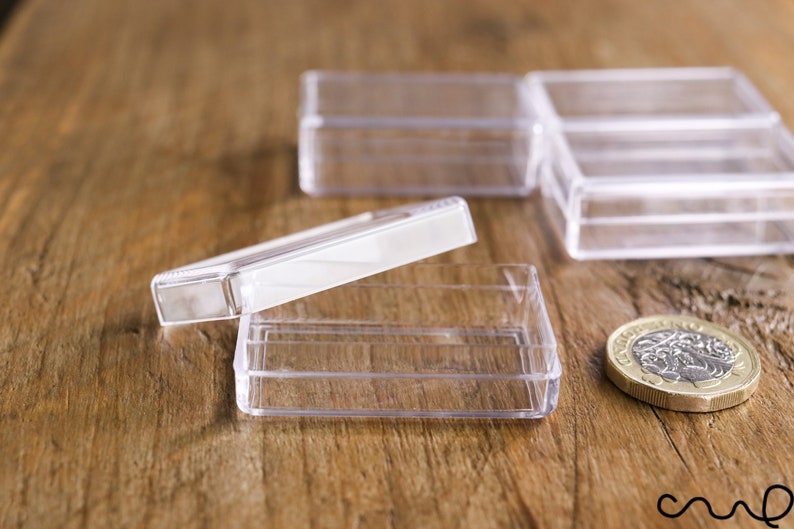 May include: Clear, rectangular plastic boxes with hinged lids, ideal for small items. One box is open, lid ajar. A British one-pound coin is next to the boxes. The boxes are on a wooden surface.