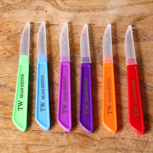 May include: Six colourful seam rippers with clear plastic blades and handles labelled "TW SEAM RIPPER".