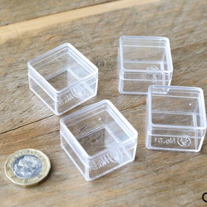 May include: Four clear plastic square boxes with lids. The boxes are approximately 1.5 inches in size. A British one pound coin is in the lower left corner of the image for scale.