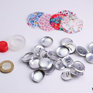 Puede incluir: Un surtido de suministros para manualidades, que incluye botones cubiertos de tela floral, botones de metal plateado, una tapa roja, un recipiente de plástico transparente y una moneda. Los botones tienen varios patrones florales.