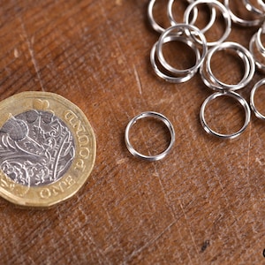 May include: A close-up of a silver jump ring next to a British one pound coin. The jump ring is about the size of a 10 pence piece. There are many more jump rings in the background.