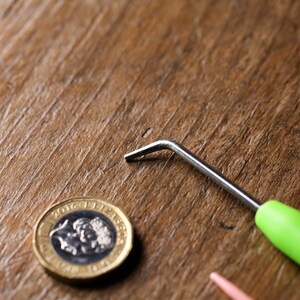 Puede incluir: Un gancho de ganchillo con mango verde y gancho de metal plateado sobre una superficie de madera, junto a una moneda brit&aacute;nica de una libra.