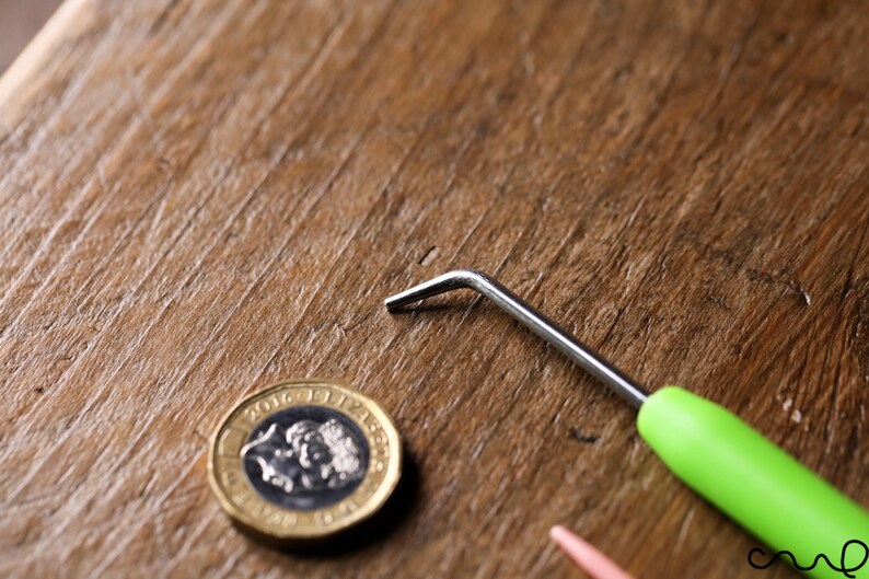 Puede incluir: Un gancho de ganchillo con mango verde y gancho de metal plateado sobre una superficie de madera, junto a una moneda brit&aacute;nica de una libra.