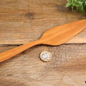 Puede incluir: Un cuchillo de mantequilla de madera con borde dentado y mango redondeado. El cuchillo es de color marrón claro y está sobre una superficie de madera. Una moneda está al lado del cuchillo.
