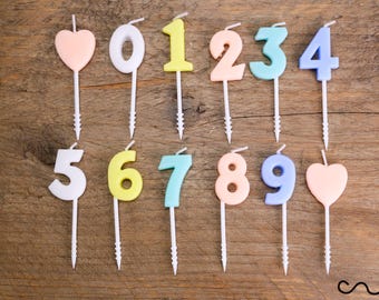 May include: A collection of colorful birthday candles shaped like numbers and hearts. The candles are in various pastel colors, including pink, yellow, blue, and peach. Each candle has a white stem with a spiral base for easy insertion into a cake.