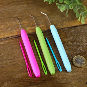 May include: Set of six colourful plastic crochet hooks with metal tips. The hooks are arranged in a row on a wooden surface. There is a coin in the background.