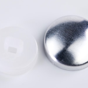 Peut inclure: Deux boutons ronds, un blanc et un argenté. Le bouton blanc a un petit trou au centre. Le bouton argenté est brillant et a une surface lisse.