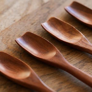 Puede incluir: Cuatro cucharas de madera con forma de pala redondeada. Las cucharas son de madera marr&oacute;n oscura y est&aacute;n dispuestas en fila sobre una superficie de madera.