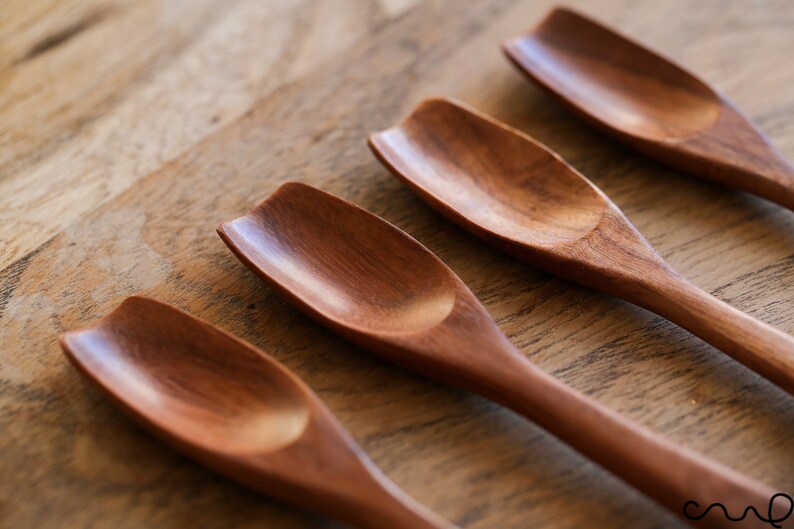 Puede incluir: Cuatro cucharas de madera con forma de pala redondeada. Las cucharas son de madera marr&oacute;n oscura y est&aacute;n dispuestas en fila sobre una superficie de madera.