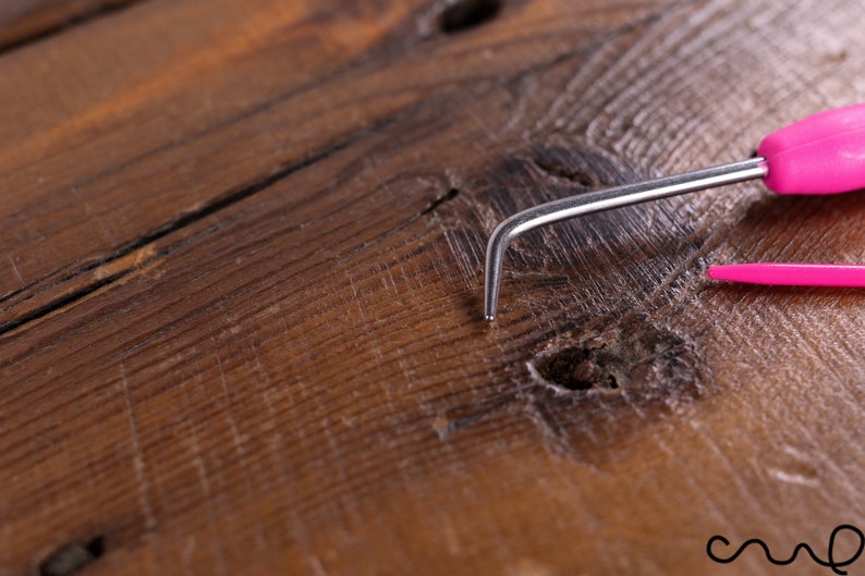 Op de afbeelding: Een close-up van een roze en zilveren haaknaald op een houten oppervlak.