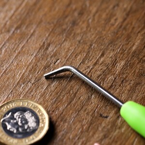 Op de afbeelding: Een haaknaald met een groene handgreep en een zilveren metalen haak op een houten oppervlak met een Britse munt van &eacute;&eacute;n pond.