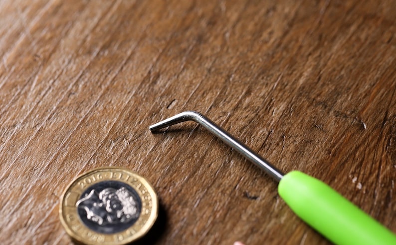 Op de afbeelding: Een haaknaald met een groene handgreep en een zilveren metalen haak op een houten oppervlak met een Britse munt van &eacute;&eacute;n pond.