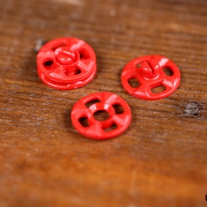 Puede incluir: Tres pequeños botones rojo brillante con diseño circular y tornillo central. Los botones son de material brillante y están sobre un fondo de madera cálida. Estos botones son probablemente para coser o manualidades.