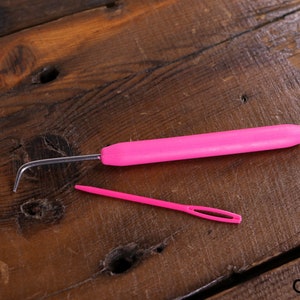 Op de afbeelding: Een roze haaknaald en een roze garennaald op een houten oppervlak.