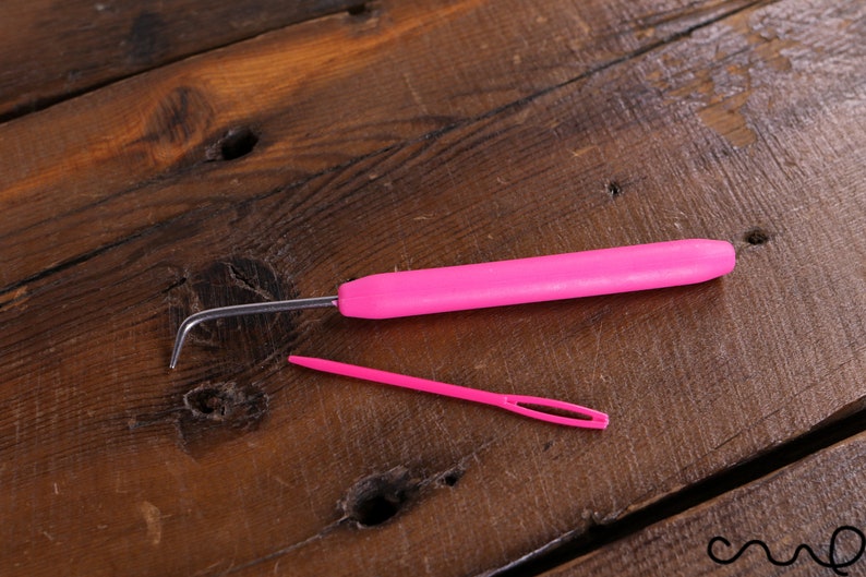 Op de afbeelding: Een roze haaknaald en een roze garennaald op een houten oppervlak.