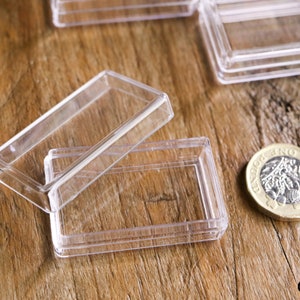 May include: Clear, rectangular plastic cases with hinged lids, ideal for small items. One case is open, revealing the interior. A British one-pound coin is next to the cases on a wooden surface.