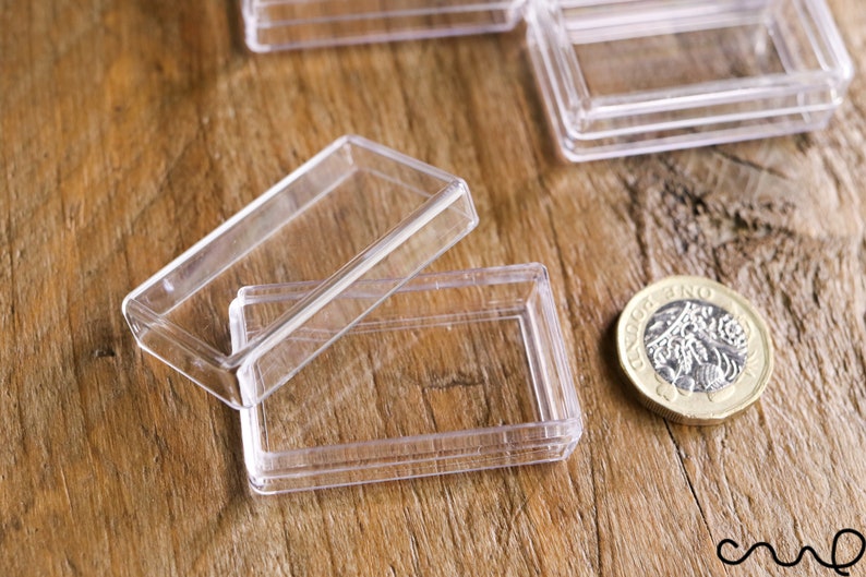 May include: Clear, rectangular plastic cases with hinged lids, ideal for small items. One case is open, revealing the interior. A British one-pound coin is next to the cases on a wooden surface.