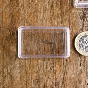 May include: A clear, rectangular plastic coin holder next to a British one-pound coin. The coin features a detailed design and is made of gold and silver-colored metal. The holder is empty and sits on a wooden surface.