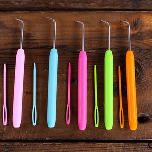 Op de afbeelding: Zeven kleurrijke haaknaalden met metalen punten, gerangschikt in een rij op een houten oppervlak. De haaknaalden zijn roze, blauw, groen, oranje en rood.