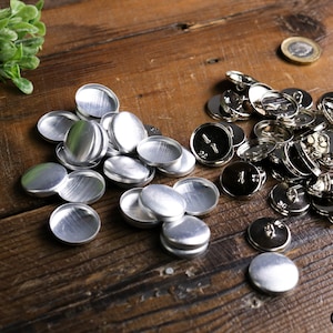May include: A close-up of two piles of silver metal buttons. One pile is of flat, round button backs, and the other pile is of complete buttons with a post and loop.