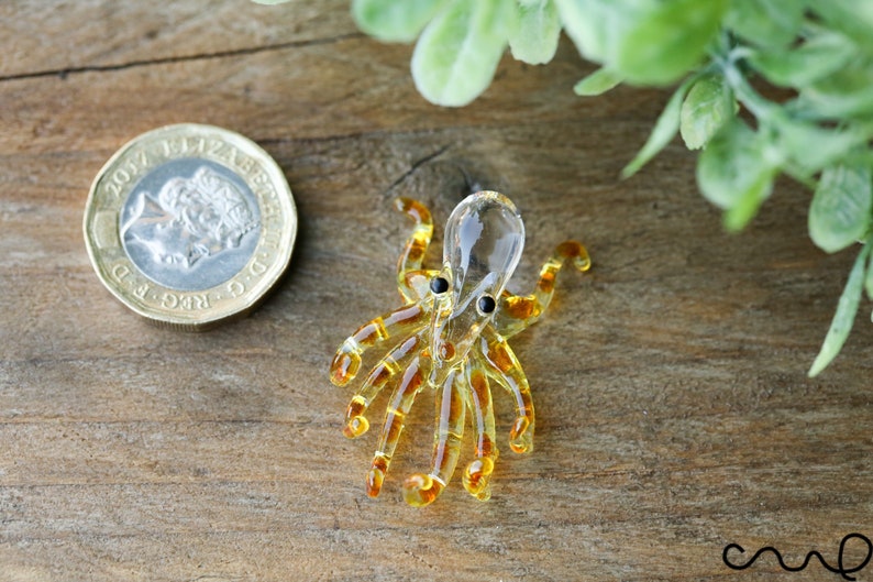Pu&ograve; includere: Una piccola statuina di polpo in vetro con un corpo trasparente e tentacoli color ambra. Il polpo ha occhi neri e si trova su una superficie di legno.