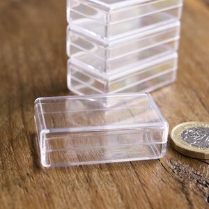 May include: Clear, rectangular plastic boxes stacked and arranged on a wooden surface. One box is in the foreground with a coin next to it. The boxes are empty and appear to be for small items.