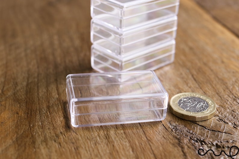 May include: Clear, rectangular plastic boxes stacked and arranged on a wooden surface. One box is in the foreground with a coin next to it. The boxes are empty and appear to be for small items.