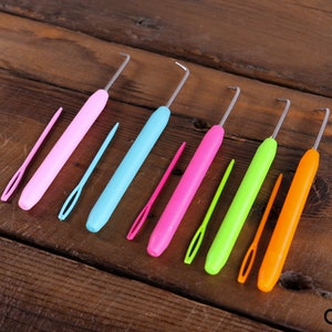 Op de afbeelding: Een set van zeven kleurrijke plastic haaknaalden met bijpassende draadtrekkers. De haaknaalden zijn gerangschikt in een rij van links naar rechts, met de grootste haaknaald links en de kleinste haaknaald rechts. De haaknaalden zijn roze, blauw, roze, groen, oranje en geel.