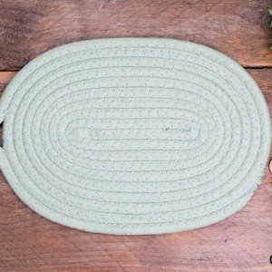 Può includere: Sottopentola ovale intrecciato verde chiaro con manico ad anello. Il sottopentola è realizzato in un materiale morbido e tessuto ed è progettato per proteggere le superfici dai piatti caldi. Il sottopentola è su una superficie di legno.