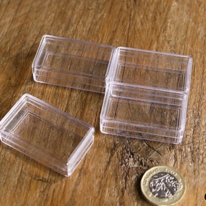 May include: Several clear, rectangular plastic boxes with hinged lids are arranged on a wooden surface. A coin is also present. The boxes appear to be empty and are likely used for storage or display of small items.