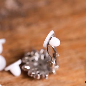 Peut inclure: Gros plan sur une boucle d'oreille argentée à clip avec un motif en cristal transparent. La boucle d'oreille est sur une surface en bois avec plusieurs dos de boucles d'oreilles en plastique blanc.