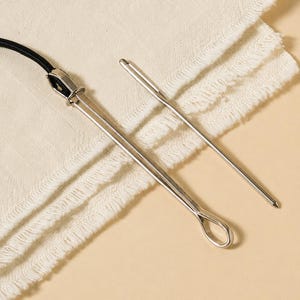 May include: Two silver-toned metal bodkins for threading elastic or cord. One bodkin features a sliding ring mechanism currently clamping a black elastic cord, while the other is a simple needle-like tool with a long eyelet for threading.