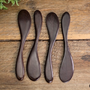 Può includere: Quattro cucchiai di legno marrone scuro disposti su una superficie di legno. I cucchiai hanno un design liscio e curvo e sono disposti in fila. I cucchiai sono di un ricco colore marrone scuro e la venatura del legno è visibile.