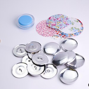 May include: A close-up of a pile of silver-colored, round, metal buttons. The buttons are arranged in a random pattern and are all the same size. There are also two pieces of fabric with floral patterns in the background.