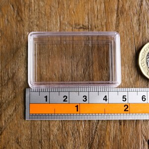 May include: A clear, rectangular plastic case with a ruler showing measurements in inches. A British one-pound coin is next to the case. The case is approximately 3 inches long.