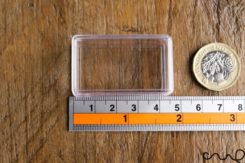 May include: A clear, rectangular plastic case with a ruler showing measurements in inches. A British one-pound coin is next to the case. The case is approximately 3 inches long.