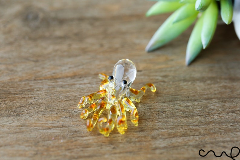 May include: A small glass octopus figurine with a clear body and brown spots. The octopus has eight tentacles and is sitting on a wooden surface.