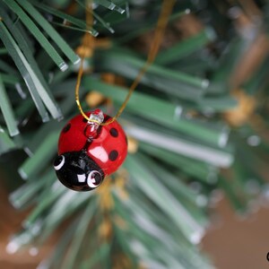 May include: A ladybird Christmas ornament with a red body and black spots hangs from a gold string. The ladybird has a black head with white eyes, set against green Christmas tree branches.